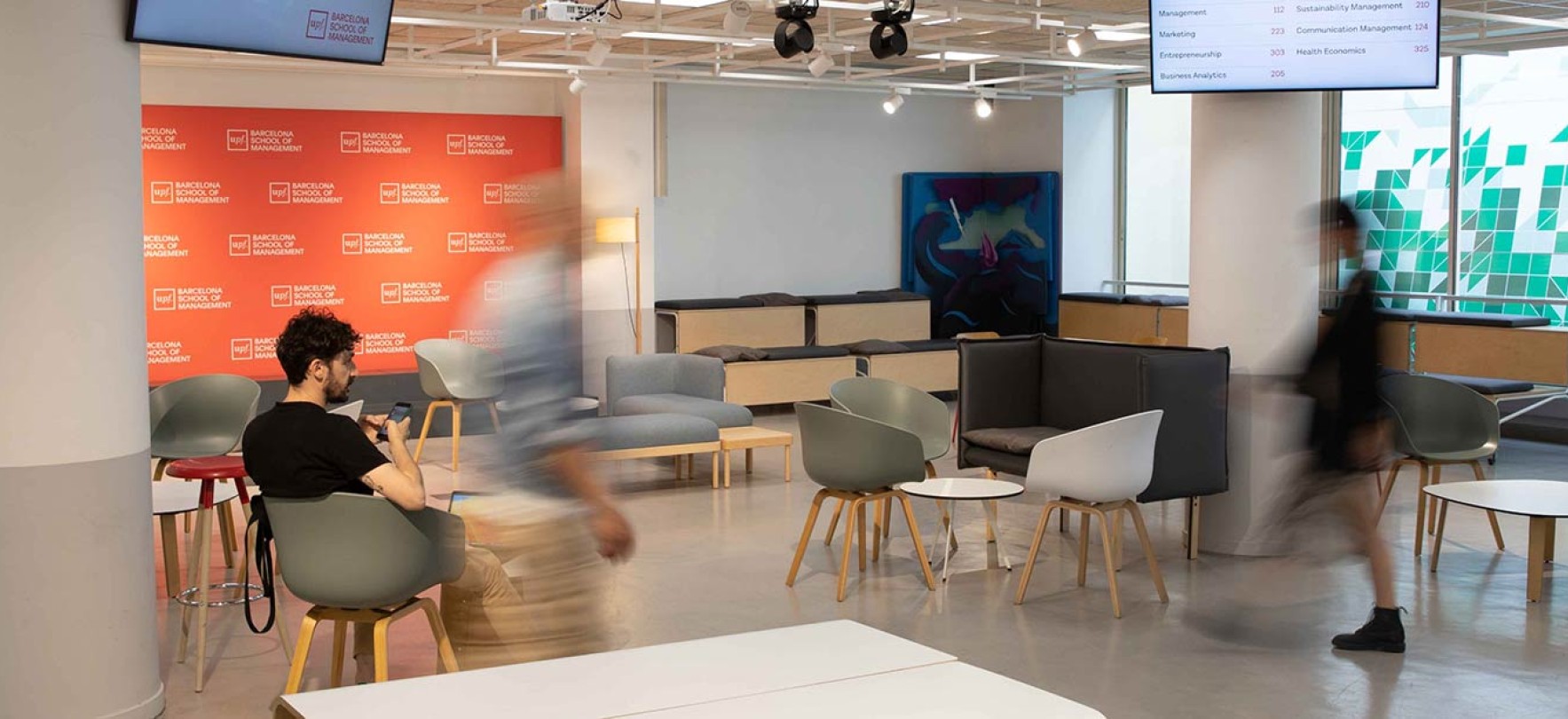 People walking through a modern university lobby with branded backdrop and seating area.